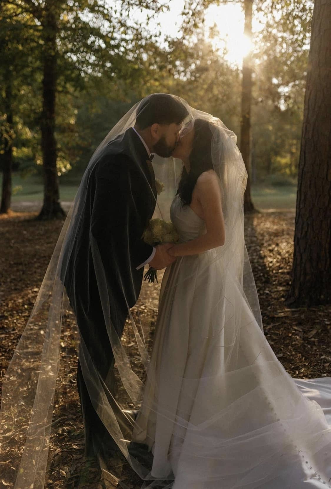 Wedding couple kissing in a forest with sunlight filtering through the trees.