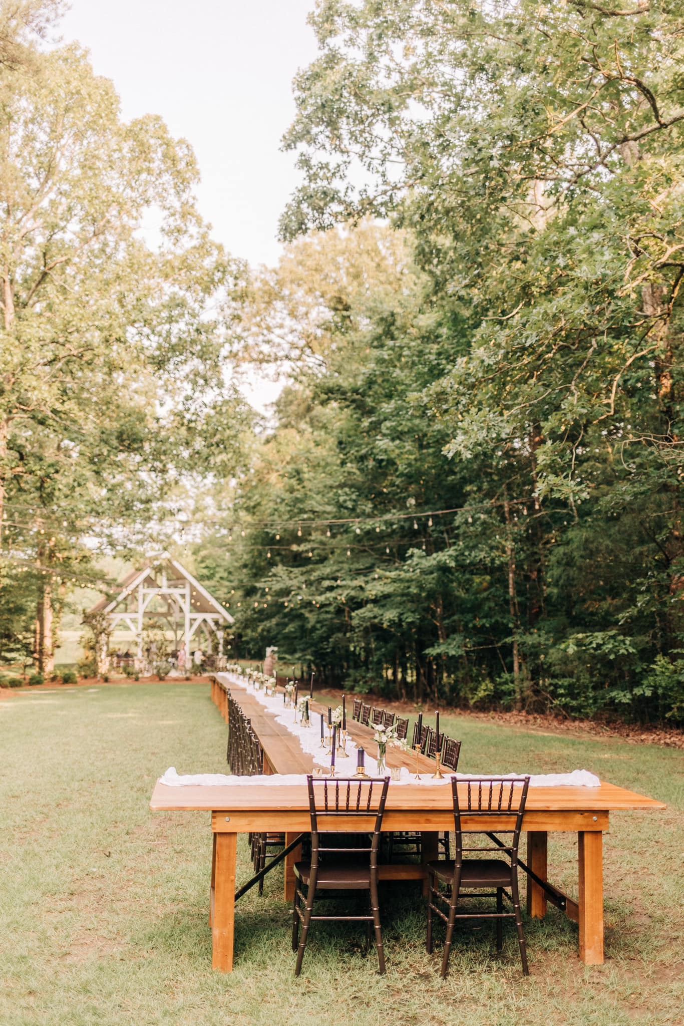 Long wooden table set for a meal in a forest setting with chairs and a covered pavilion in the background.