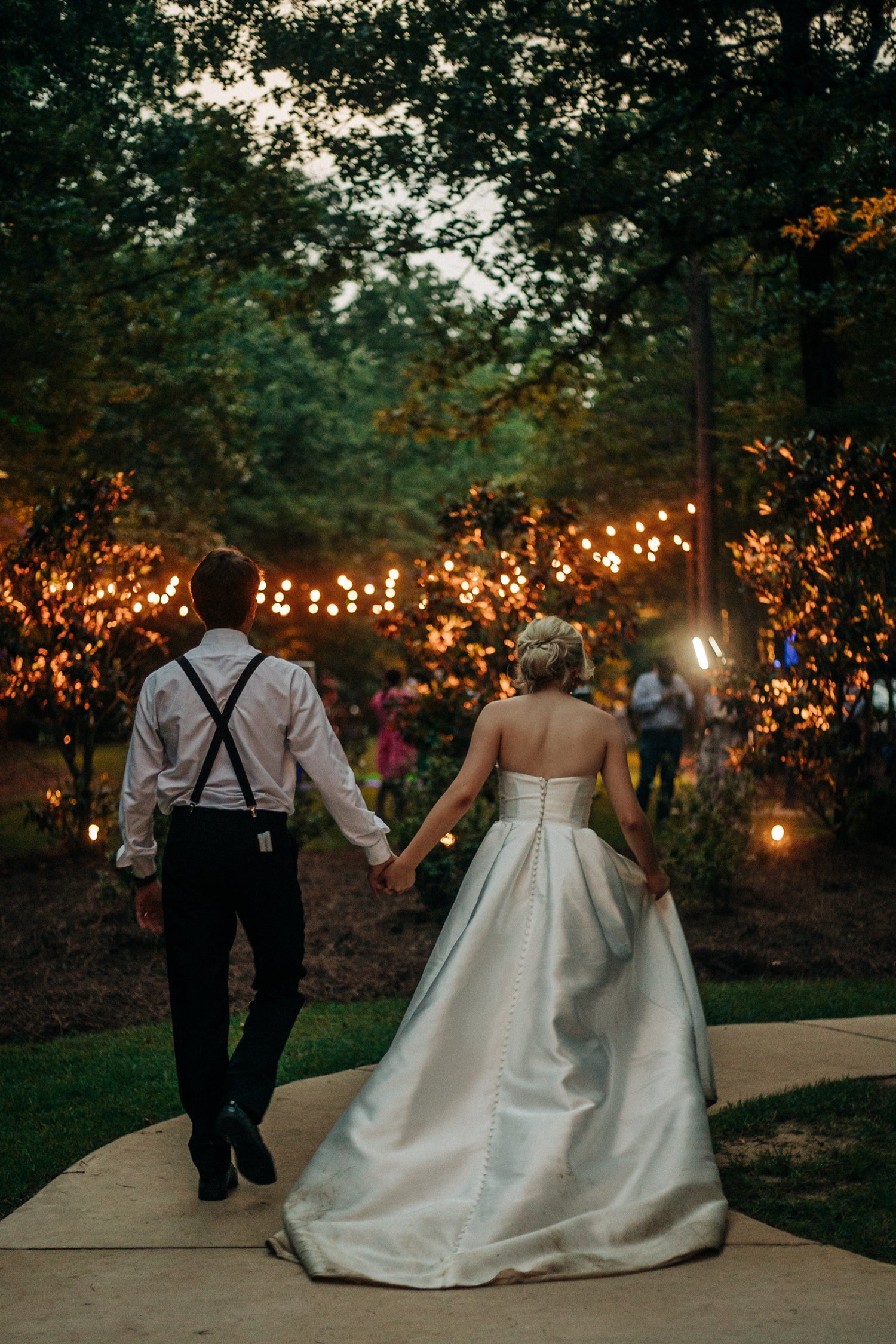 bride and groom holding hands in a garden with string lights at night at Brookwood Venue