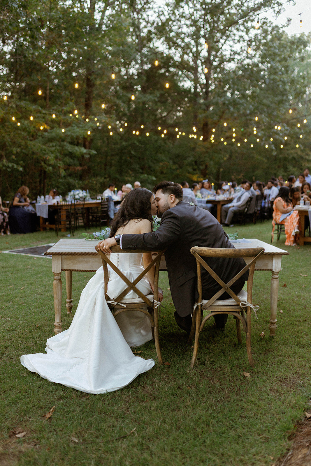 Couple sharing a kiss at an outdoor wedding reception with string lights and guests in the background.