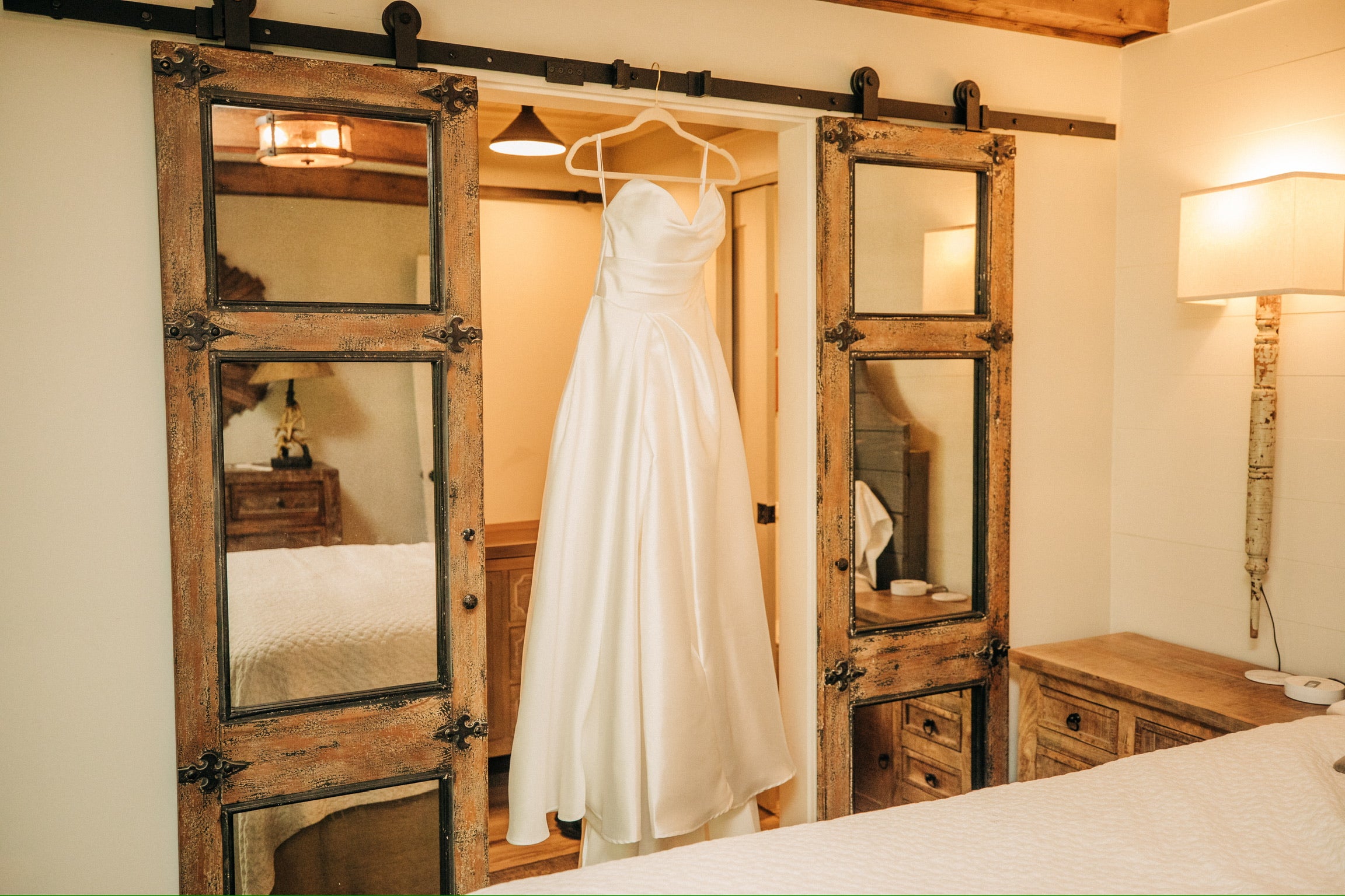 White wedding dress hanging on a wooden hanger inside a rustic room with mirrors.